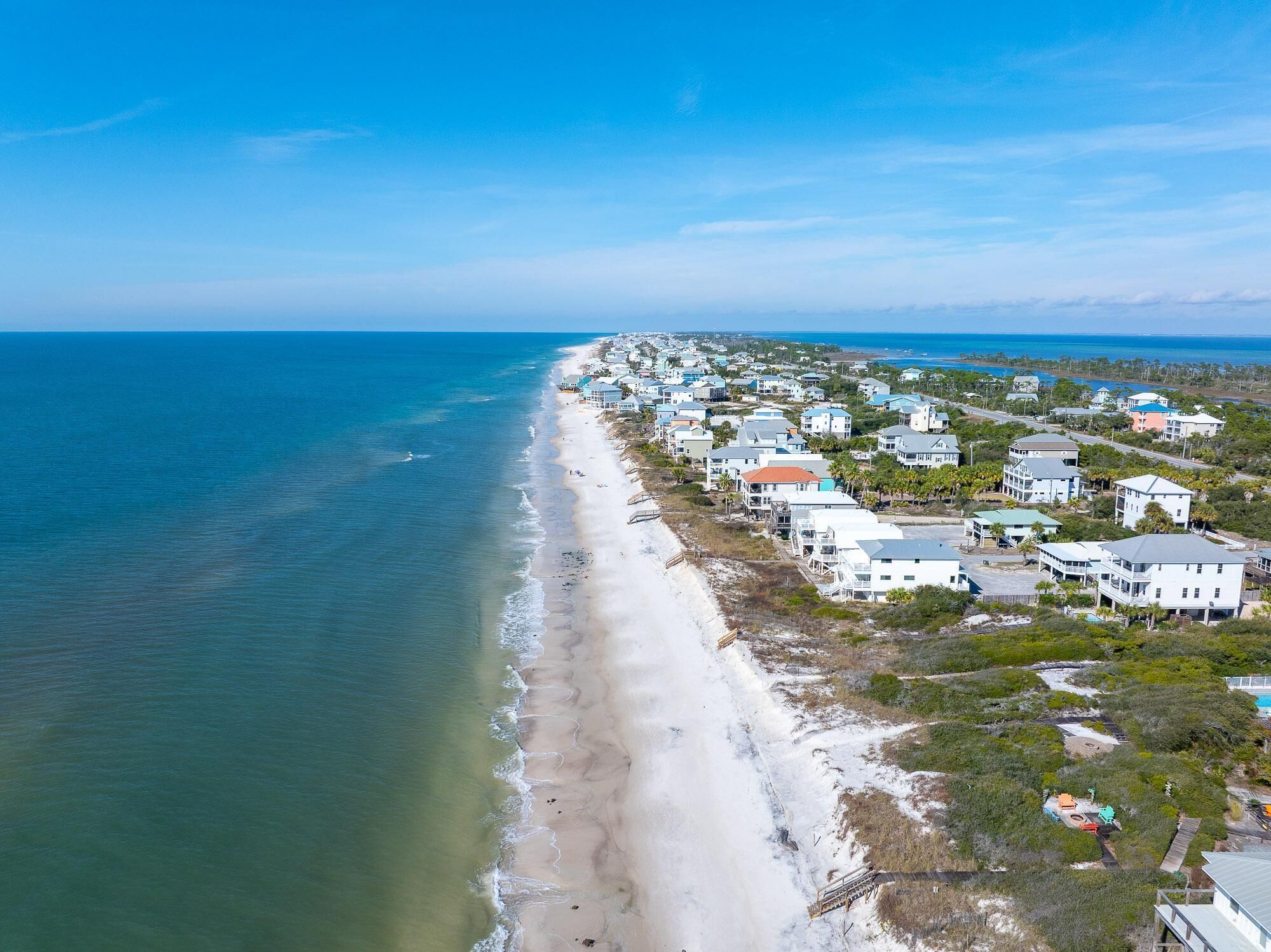 An aerial view of a coastal residential area with houses along the beachfront, a clear sky, and calm sea.