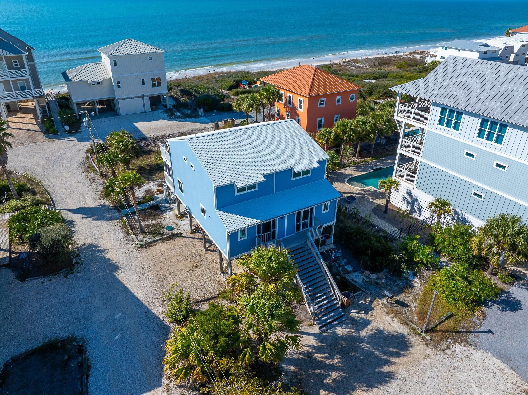 A coastal property with multiple buildings, including a prominent blue house with a staircase leading to a deck, surrounded by palm trees and a beach view.