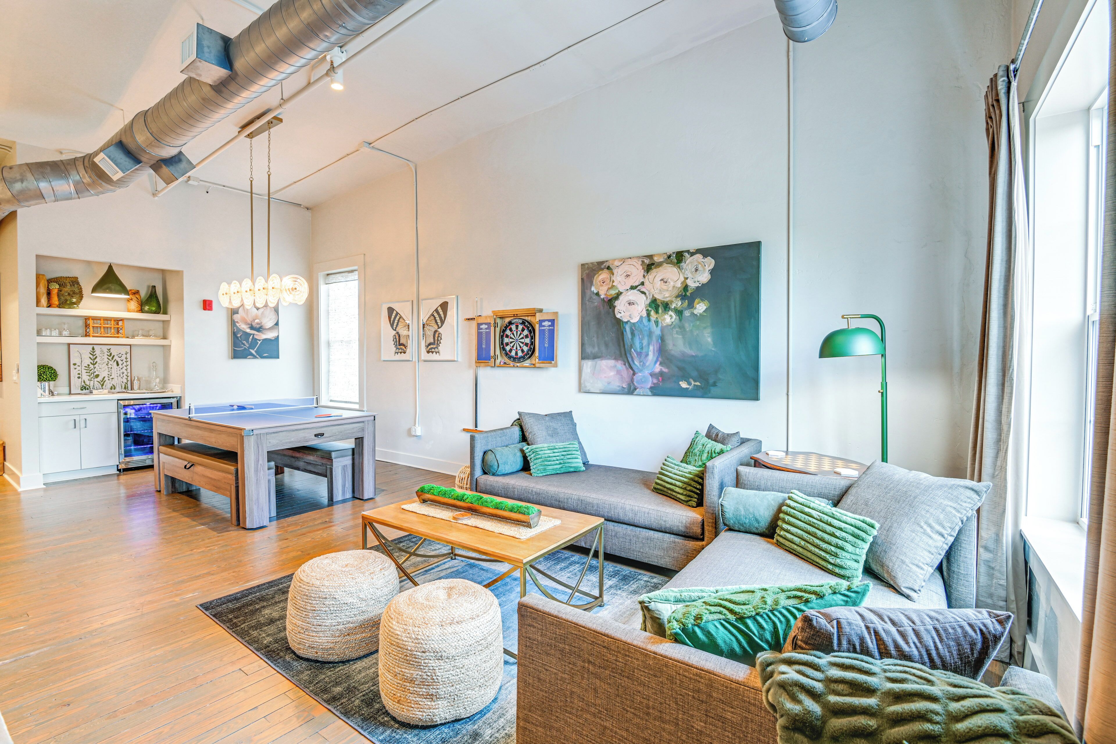 A spacious living area with a sofa set, a coffee table, and a dartboard. There is a painting on the wall and a green lamp.
