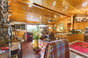Interior rústico de una cabaña con chimenea de piedra, techo de madera y una zona de estar acogedora con una mesa de comedor, sillas y un tapete rojo.