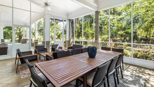 Un patio cubierto con una mesa de madera y sillas de mimbre, ventanas amplias que dan a zonas verdes.