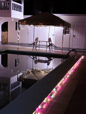 A pool area with a canopy and string lights, surrounded by a white building with balconies. - Patch and Porch (Port Antonio)