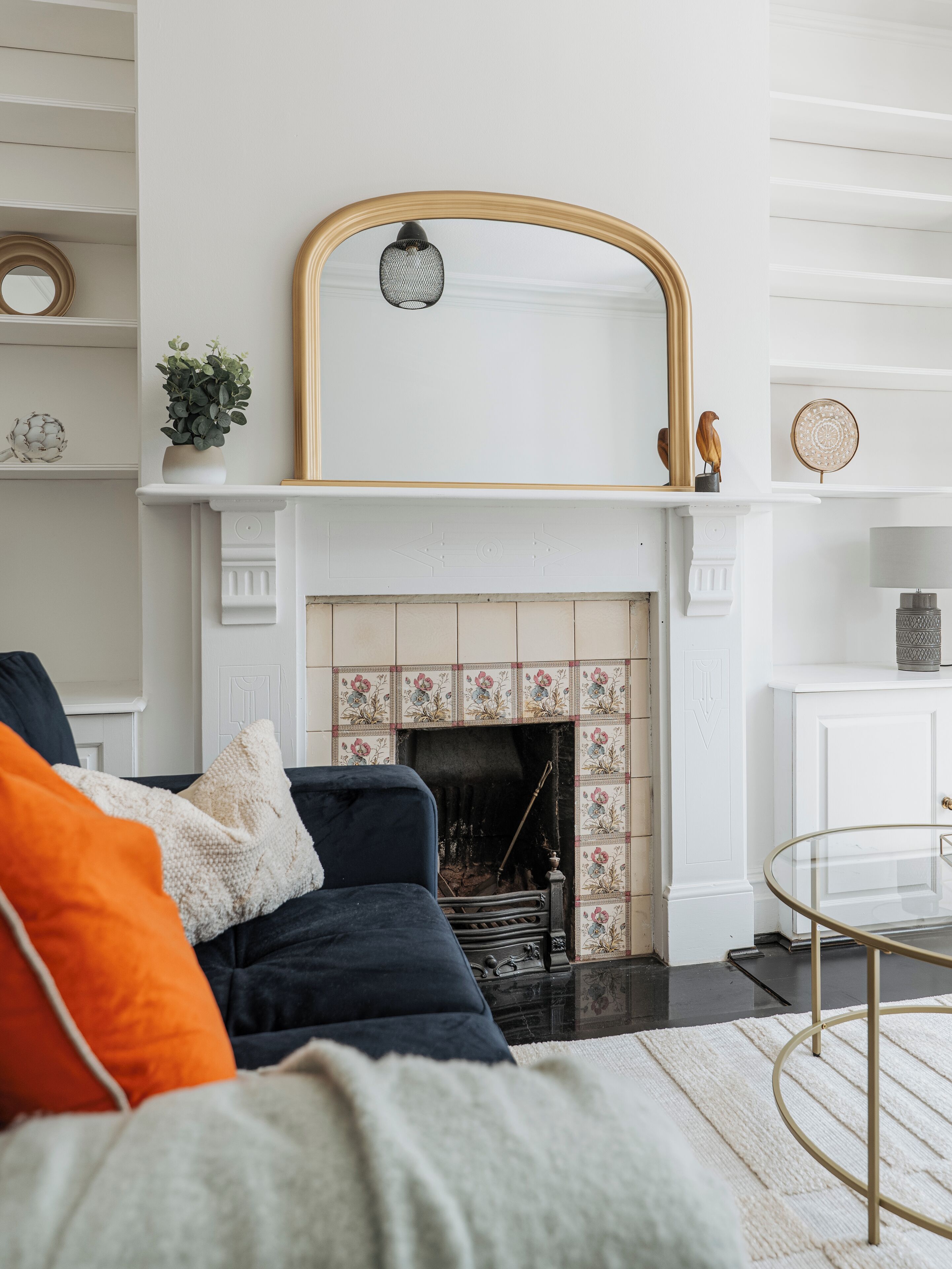 Un salon avec une cheminée, un canapé avec des coussins, une table basse en verre et un miroir décoratif au-dessus de la cheminée.