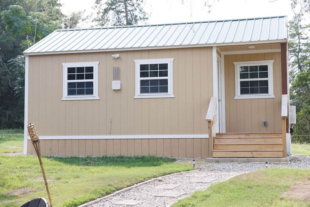 A single-story, beige wooden building with two windows and a door, surrounded by a gravel path and grass.