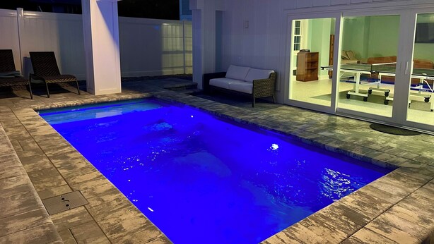 Una piscina con luz azul, un sofá exterior blanco y una mesa para jugar al ping pong.