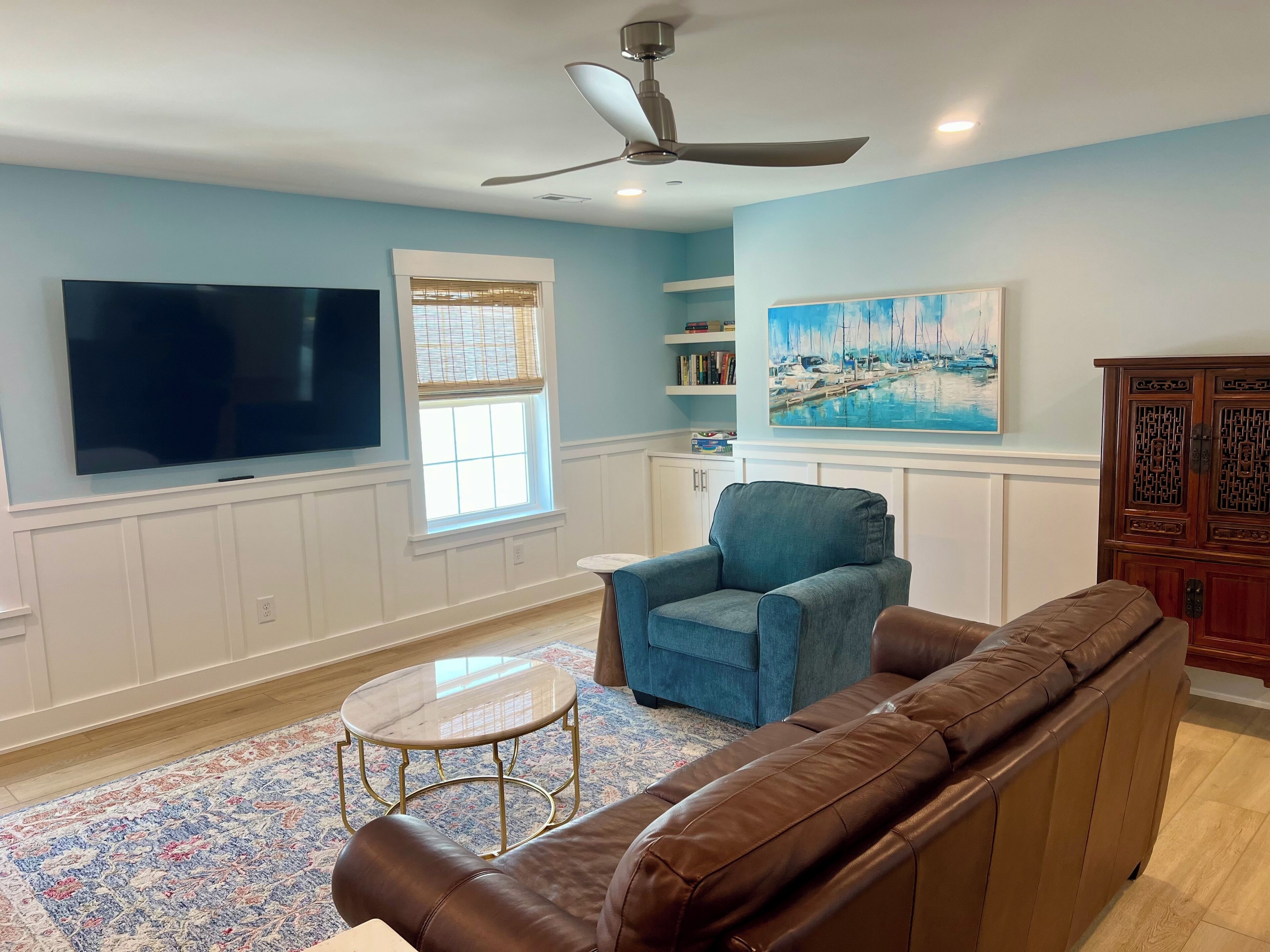 A living room with a flat-screen TV, a ceiling fan, a blue armchair, a brown leather sofa, a small round coffee table, and a painting of a marina.
