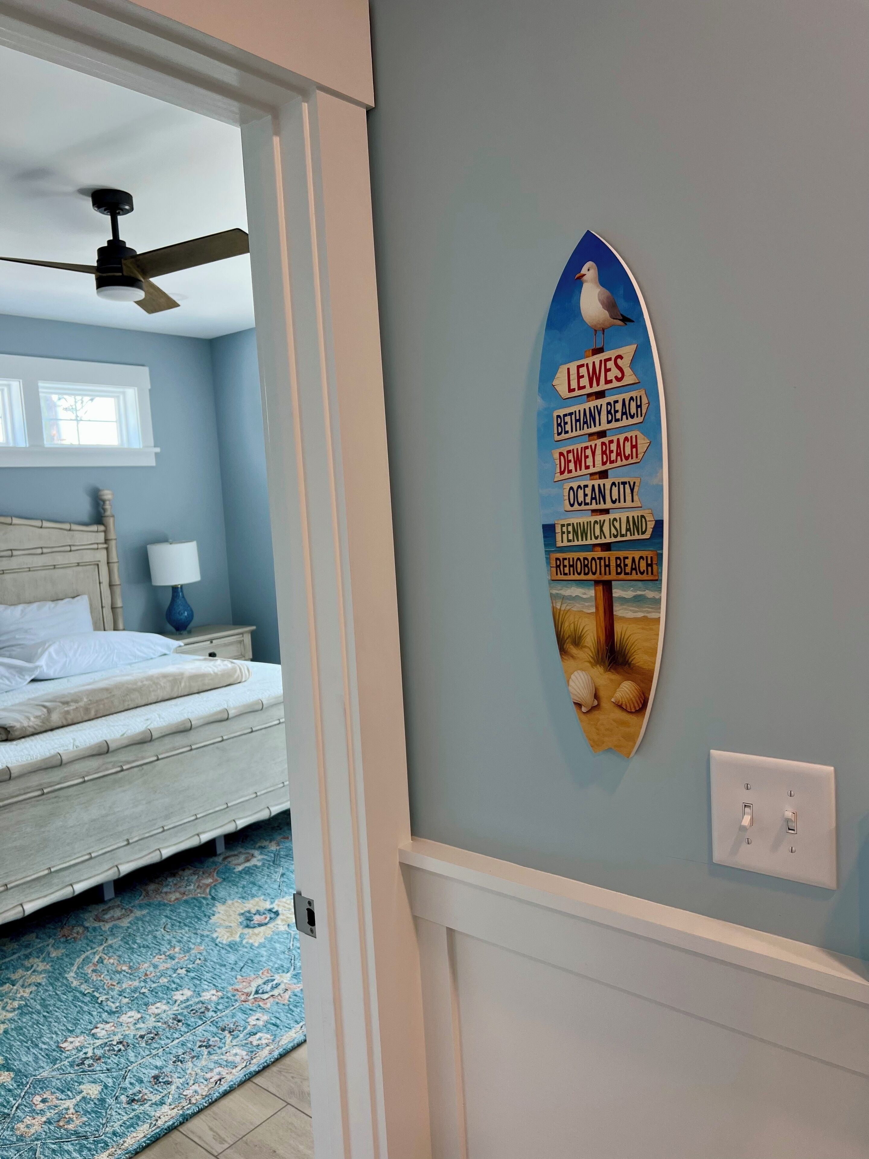 A bedroom with a bed, a nightstand, a lamp, a ceiling fan, and a wall with a beach-themed sign listing various beaches.