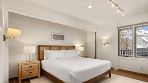 A bedroom with a wooden bed, two bedside tables with lamps, a window with a view of snow-covered mountains, and a framed picture on the wall. - Lichenhearth 29 (Snowmass Village)