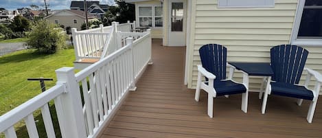 Una terraza de madera con barandilla blanca, un espacio de estar cubierto con dos sillas y una casa al fondo.