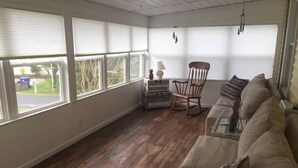 Una habitación amplia con un ventanal grande, suelo de madera, un sofá color beige, un sillón mecedora y una mesita con lámpara.