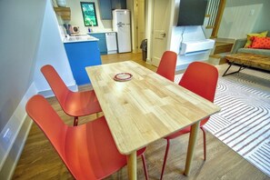 A dining area with a wooden table and red chairs, a kitchen in the background, and a living area with a sofa and patterned rug.