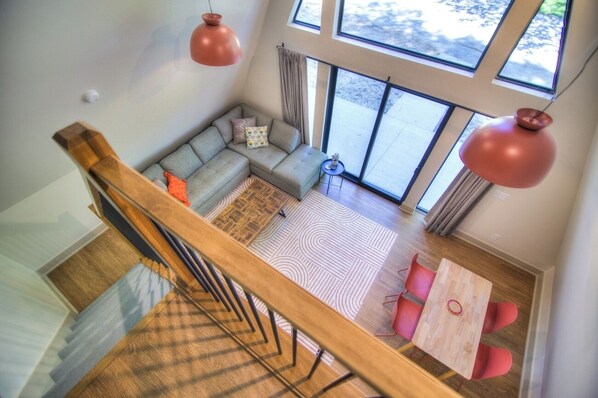 A modern living room with a sofa, a small table, and chairs. There are large windows and a staircase.
