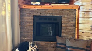 Un saló acollidor amb una llar de pedra, un televisor de pantalla plana muntat a la paret, i un gos relaxant-se sobre un coixí.