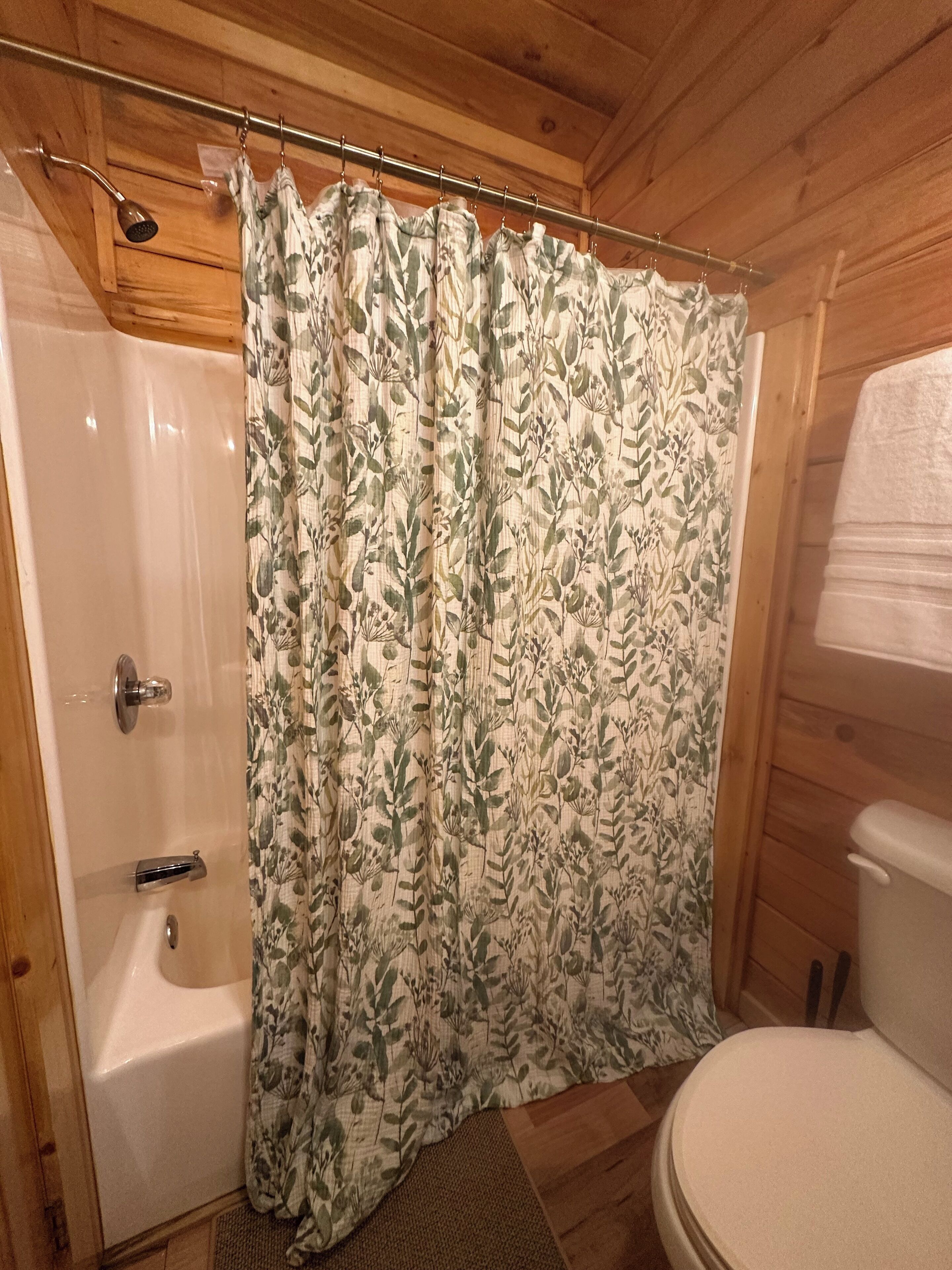 A wooden cabin bathroom with a shower, toilet, and curtain with a leaf pattern.