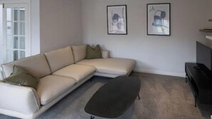 Ein Wohnzimmer mit einem beigefarbenen Ecksofa, einem dunklen Ottomanen und zwei gerahmten Kunstwerken an der Wand.