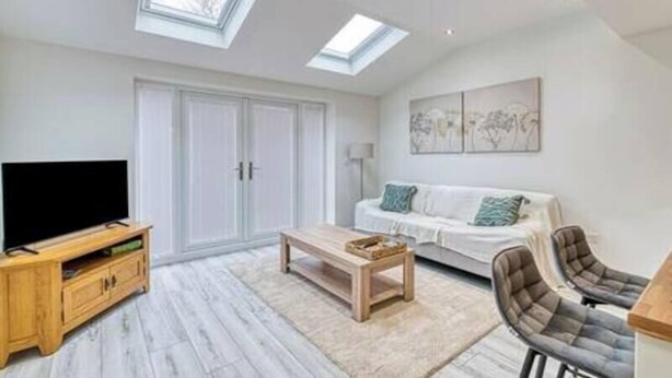 A modern living room with a flat-screen TV, a wooden coffee table, a sofa, and a chair. There are skylights on the ceiling, a large window, and artwork on the wall. - Middlecroft Annex by Guestz (Sutton)