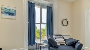 Ein Zimmer mit einer schwarzen Ledersofa, einem kleinen runden Tisch, einem Fenster mit blauen Vorhängen und einem achteckigen Spiegel im Rahmen an der Wand.
