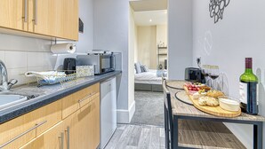 Eine kompakte Küche mit modernen Geräten, ein Essbereich mit Holztisch und ein kleines Schlafzimmer, das durch eine offene Tür sichtbar ist.