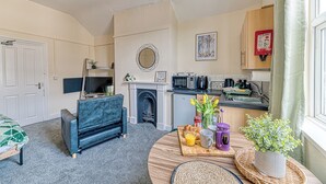 A compact living space with a sofa, a small dining table with breakfast items, a fireplace, a kitchen area, and a window with curtains.