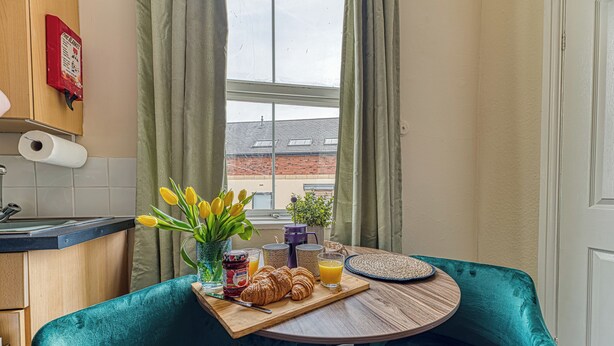 A breakfast setup with a tray of croissants, orange juice, and jam, a vase with yellow flowers, and a view of a brick building through a window.