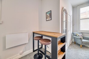 Una habitación con una mesa de madera y dos taburetes, una puerta con panel de vidrio, un cuadro en la pared y una ventana con persianas.