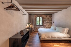 A bedroom with a bed, a wooden dresser, a hanging coat rack, a stone wall, and a window with curtains.