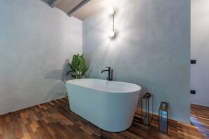 A modern bathroom with a freestanding bathtub, wooden flooring, and a wall-mounted light fixture.
