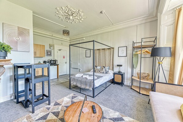 A modern hotel room with a four-poster bed, a round wooden coffee table, a small side table, a desk with a computer, a bookshelf, and a large mirror. - Chester Heights by Guestz (Chester)