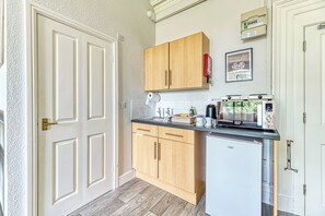 A compact kitchen with wooden cabinets, a countertop with a coffee maker, a small refrigerator, and a window with a view of greenery. - Chester Heights by Guestz (Chester)