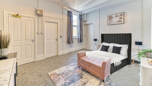 A bedroom with a bed, bedside tables, a nightstand, a lamp, a window with curtains, a door, and a ceiling light fixture. - Chester Heights by Guestz (Chester)