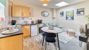 A compact kitchen with wooden cabinets, a microwave, and a small refrigerator. There is a dining area with a round table and chairs, and a sofa. - Chester Heights by Guestz (Chester)