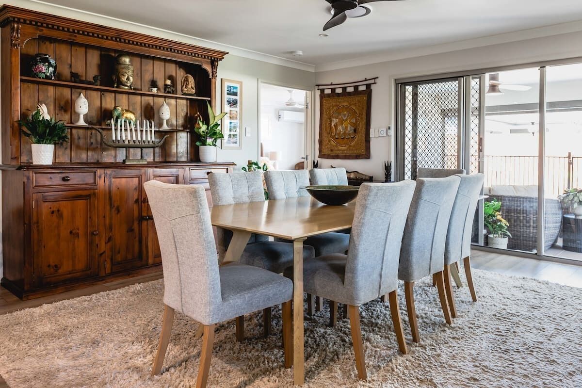 A dining area with a wooden table and grey upholstered chairs, a wooden cabinet with decorative items, and a large window with a view of the outdoors.