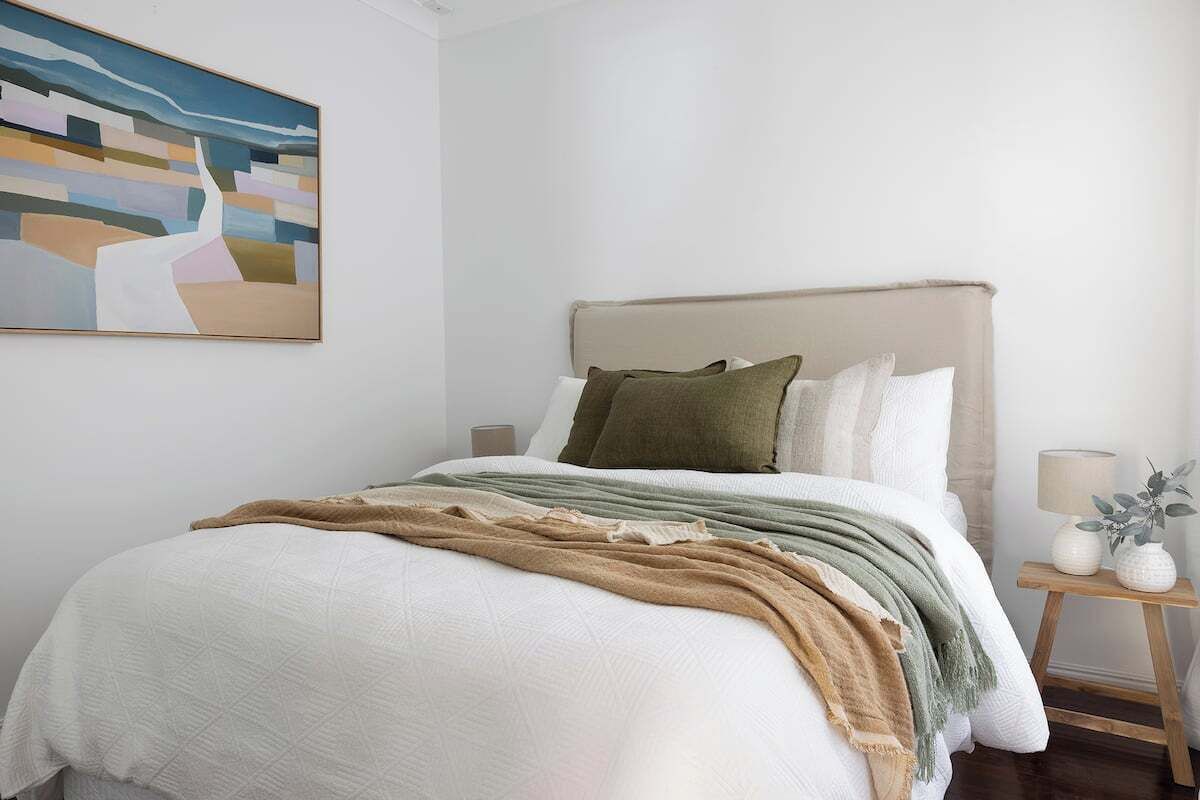 A neatly made bed with a beige headboard, a white quilt, and a green throw blanket. There is a small bedside table with a lamp and a vase of flowers. A painting is hung on the wall above the bed.