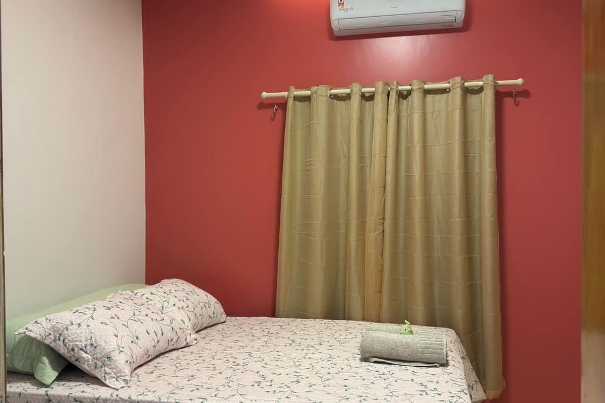 A small, single-bed room with a red wall, a beige curtain, and a white air conditioning unit above the bed.