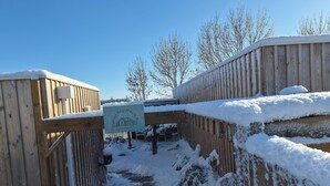 Una valla de madera con un cartel, suelo cubierto de nieve y árboles sin hojas al fondo.