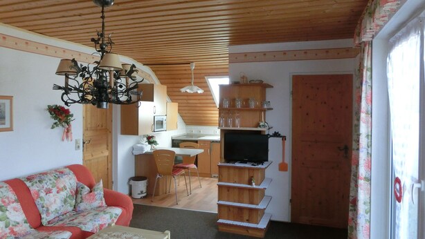 Una sala de estar acogedora con un sofá floral, una mesa de comedor de madera y una cocina al fondo.