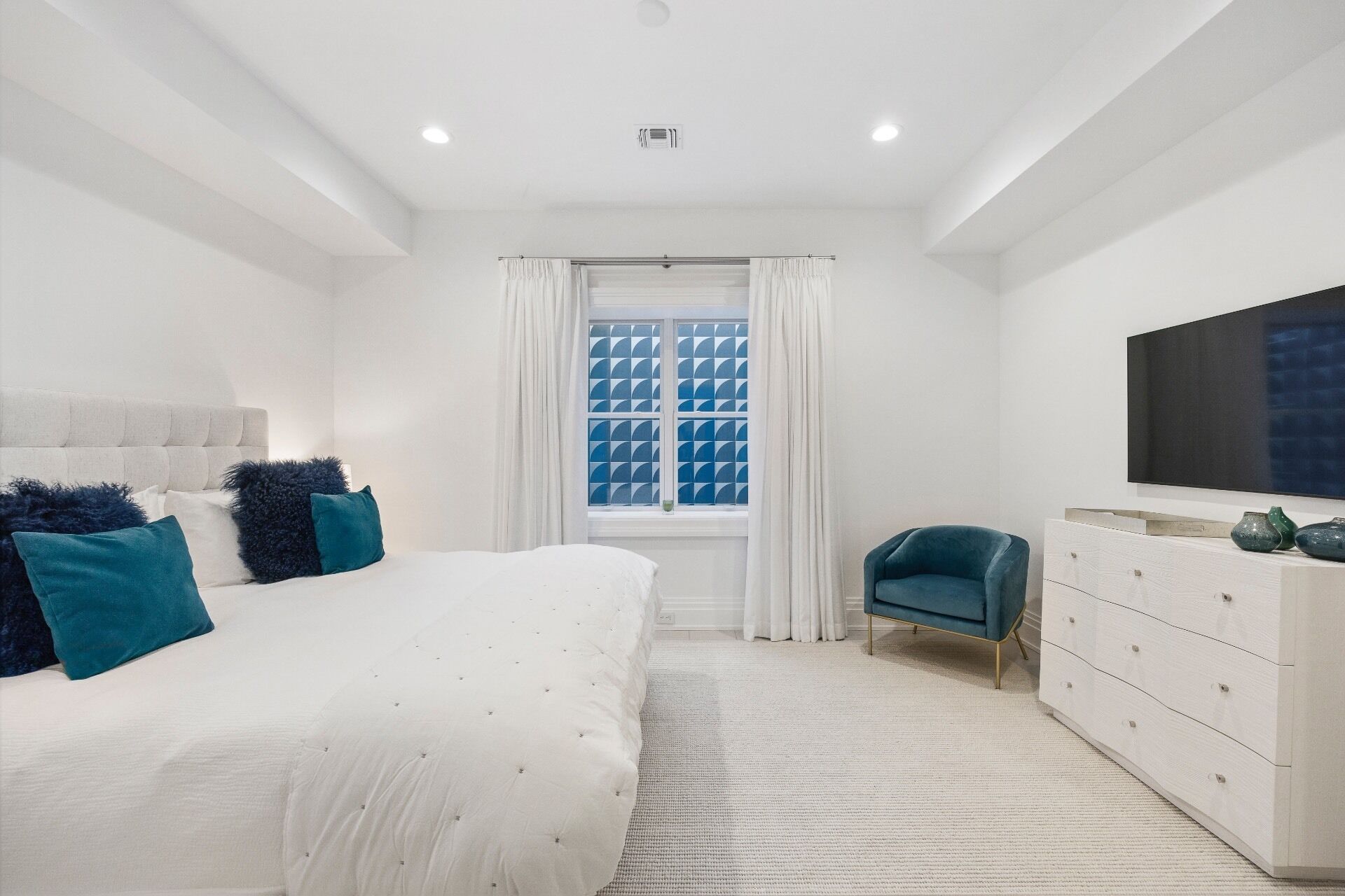 Quarto moderno com uma cama grande, uma televisão, uma cômoda e uma janela com cortinas estampadas.
