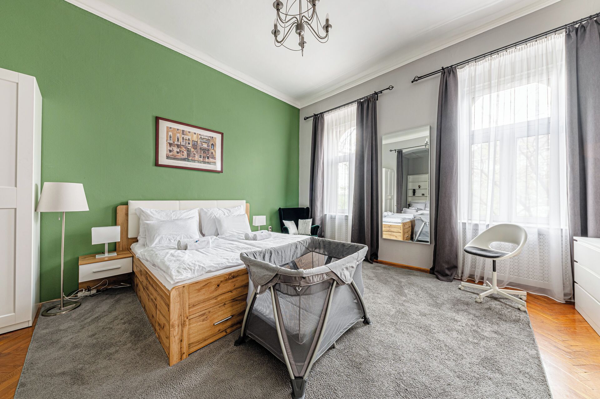 A bedroom with a green wall, a wooden bed, a grey baby crib, a white chair, and a large mirror.