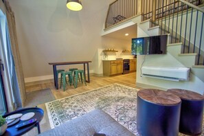 Amplia sala de estar con escalera, una mesa de comedor con taburetes, una cocina al fondo y un ventanal grande.