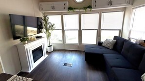Una sala de estar con chimenea, un sofá, un televisor montado en la pared y ventanas grandes con persianas.