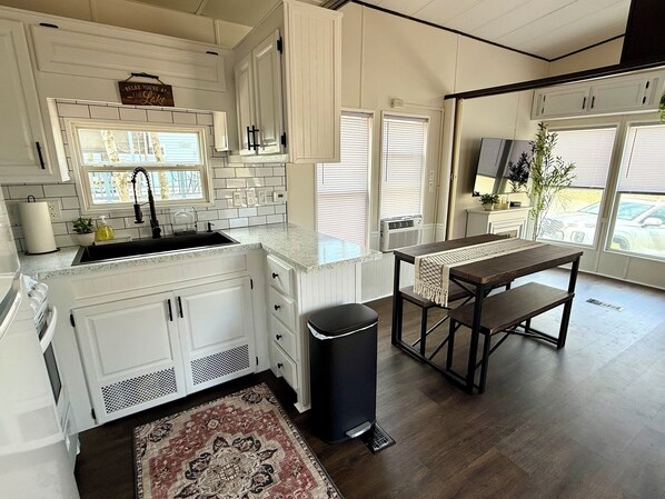 A modern kitchen with white cabinetry, a black trash bin, a wooden dining table, and a patterned rug. - Charming tiny home in wonderful private community, Eagle Lake (Eagle Lake)