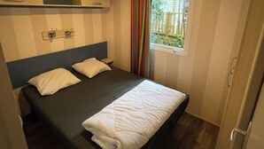A small, single-bed room with a wooden headboard, a window with curtains, and a built-in shelf above the bed. - 3-bedroom mobile home with pool (Camiers)