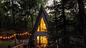 A wooden cabin with a triangular roof, surrounded by trees. It features an outdoor seating area with a table and chairs, and string lights. - Prunella № 3, Prunella № 3 - A-Frame Cabin Getaway - Design, Comfort & Nature (Thorne)