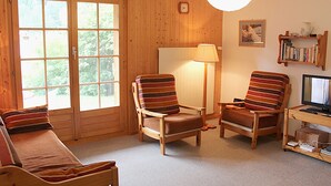 Tunnelmallinen huone, jossa on puuseinät, suuri ikkuna, kaksi raidallista tuolia, sohva, tietokoneella varustettu työpöytä ja kirjahylly.