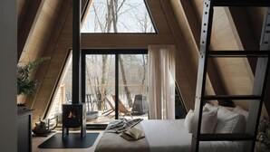 Ein gemütliches Schlafzimmer unter dem Dach mit einem großen Fenster, einem Schlafplatz in einem hölzernen Hochbett, einem Kamin und einer Leitern.