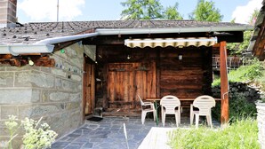 Il s’agit d’un bâtiment rustique en bois, doté d’un mur en pierre et d’un toit en tuiles. On y trouve deux chaises en plastique et une table à l’extérieur.