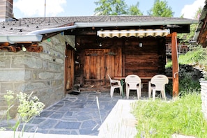 A rustic wooden building with a stone wall and a tiled roof. There are two plastic chairs and a table outside. - Mayen Motau - Retour à l'essentiel | La Forclaz (La Forclaz)