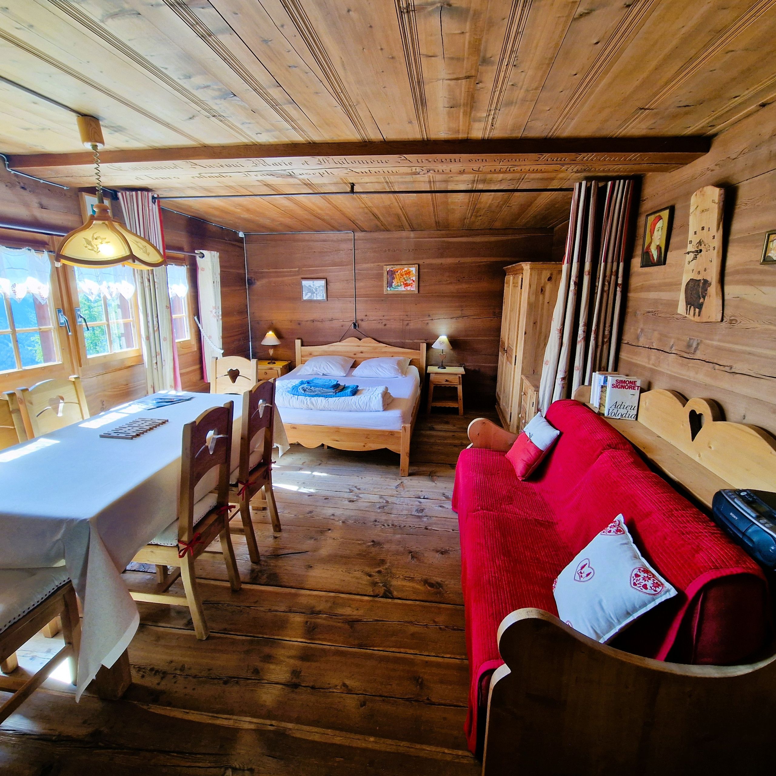 A wooden cabin interior with a bed, a red sofa, a dining table, and a chandelier.