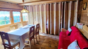 A wooden cabin with a dining area featuring a table and chairs, a red sofa, and a window with curtains. - Mayen Motau - Retour à l'essentiel | La Forclaz (La Forclaz)