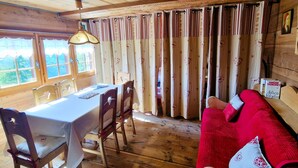Une cabane en bois comprenant un coin repas avec une table et des chaises, un canapé rouge et une fenêtre avec des rideaux.